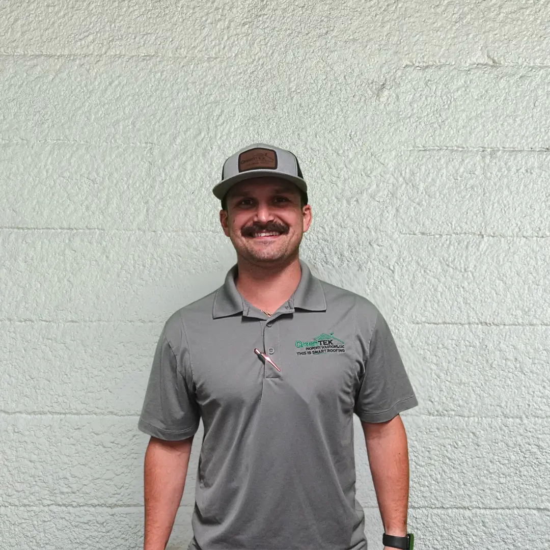 A man with a mustache wearing a gray hat and a gray polo shirt stands smiling in front of a textured light-colored wall.