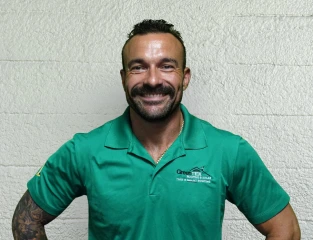 A smiling man with short dark hair and a beard wears a green polo shirt with a company logo, standing against a textured light-colored wall with his hands on his hips.
