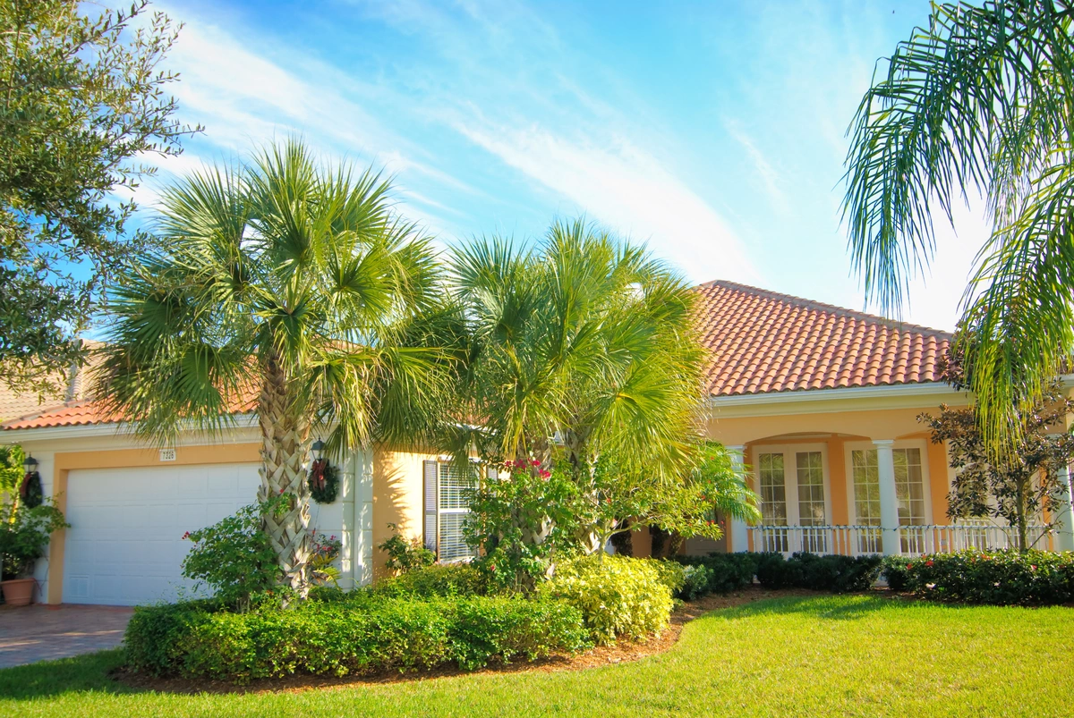A single-story house with a classic red-tiled roof, highlighting the charm of tile roofing when considering metal roof vs tile roof in Florida, surrounded by lush green palm trees and shrubs under a blue sky. A white garage door sits on the left side.
