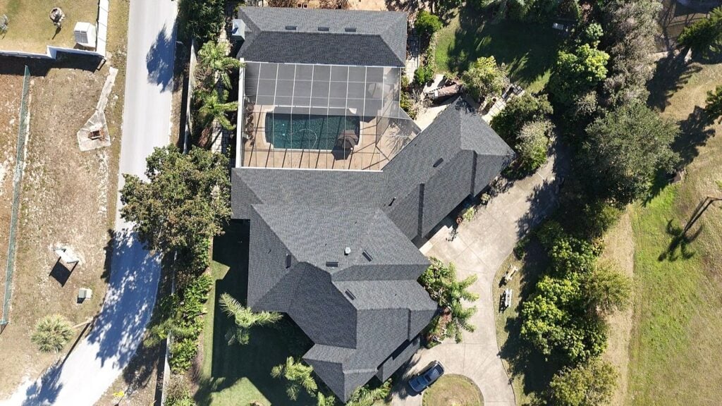Aerial view of a large house with dark roofing, a screened-in backyard pool, curved driveway, and surrounding green trees and lawn. A street borders the property on the left side.