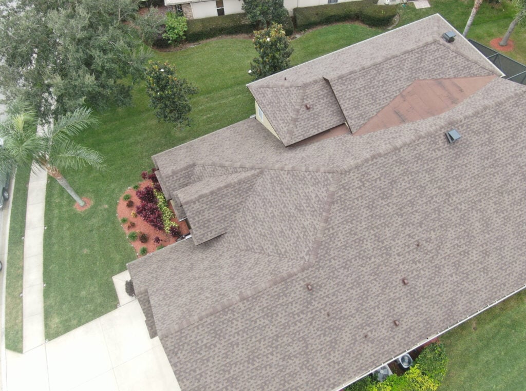 Aerial view of a house with a brown shingle roof, surrounded by green grass, trees, and landscaped shrubs near a driveway.