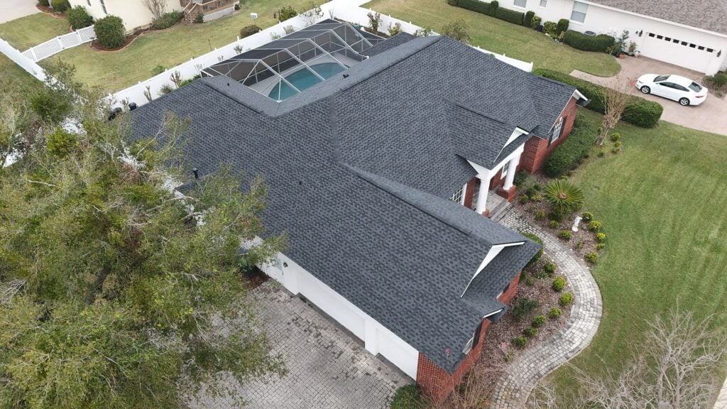 Aerial view of a house with a dark gray shingle roof, red brick and white exterior, attached garage, landscaped yard, and a screened-in backyard pool. Nearby homes, trees, and a white car are also visible.