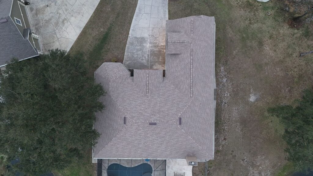 Aerial view of a single-story house with a light gray roof, a driveway leading to a garage, and a small pool area in the backyard. Trees surround part of the house, and the yard appears mostly grassy.