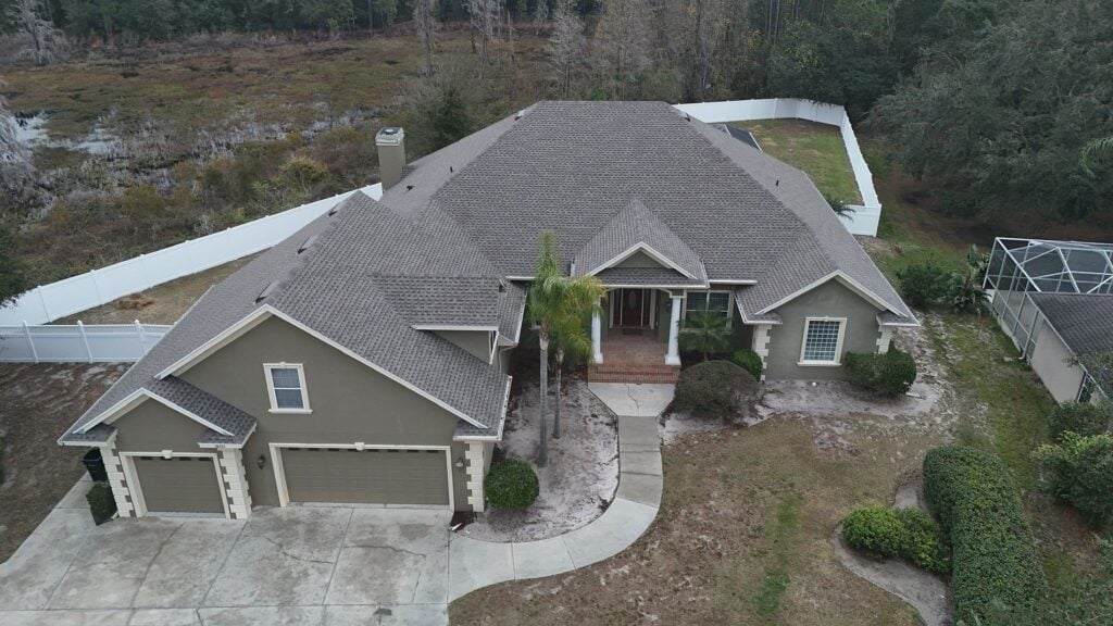 Aerial view of a large, single-story house with a gray roof, three-car garage, driveway, and white fenced backyard, surrounded by trees and a grassy yard.