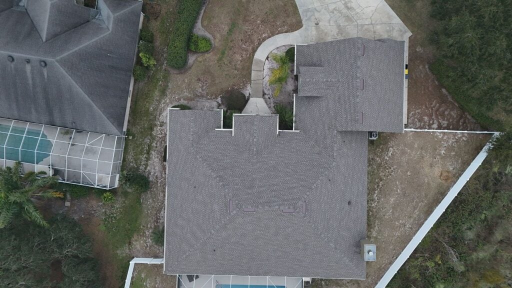 Aerial view of a house with a gray roof, surrounded by a driveway, yard, white fence, and neighboring homes; one neighbor has a screened pool area.