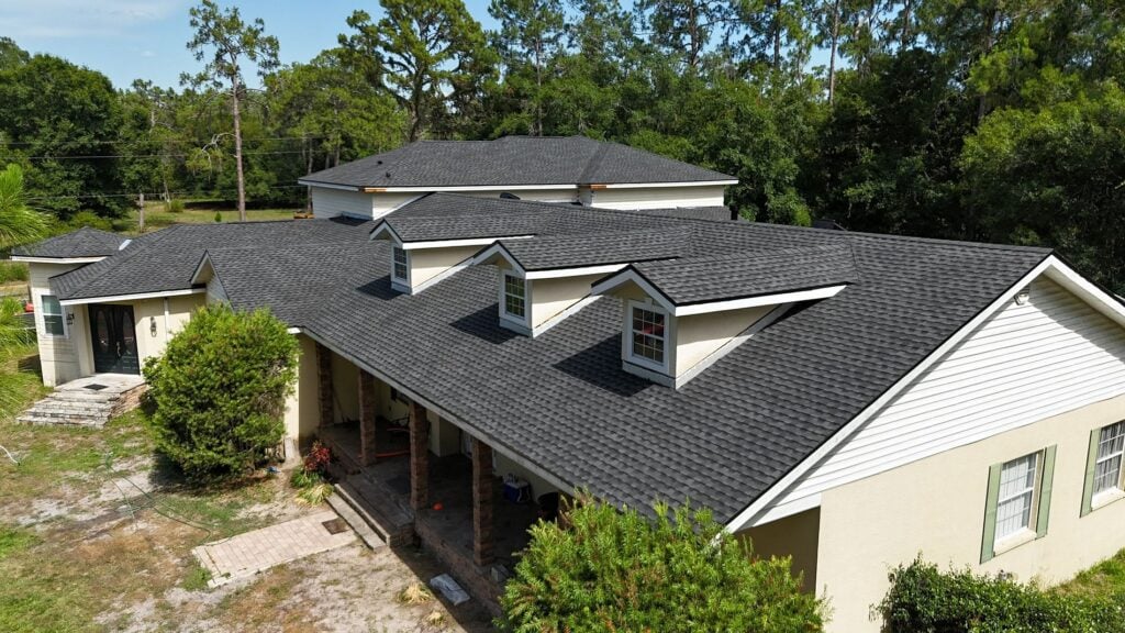 A beige house with a dark gray shingle roof featuring multiple dormer windows, surrounded by trees and greenery under a clear sky.