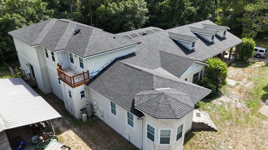 A large, light-colored two-story house with multiple rooflines and dormer windows, surrounded by trees. There is a small balcony, a covered carport, and various items on the ground near the building.