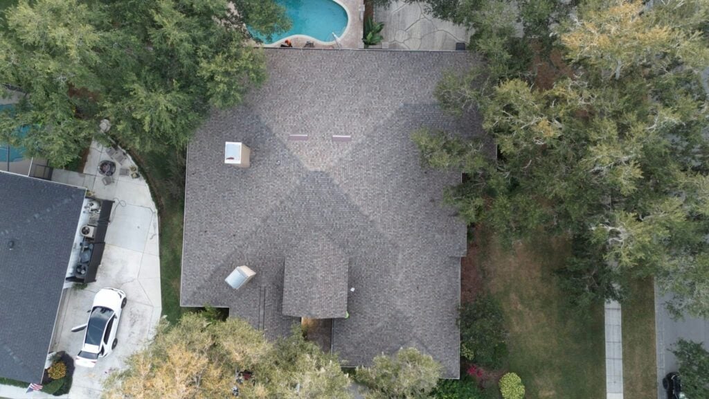 Aerial view of a house surrounded by trees, with a gray shingle roof and a driveway with a white car parked. A swimming pool and patio area are visible in the backyard.