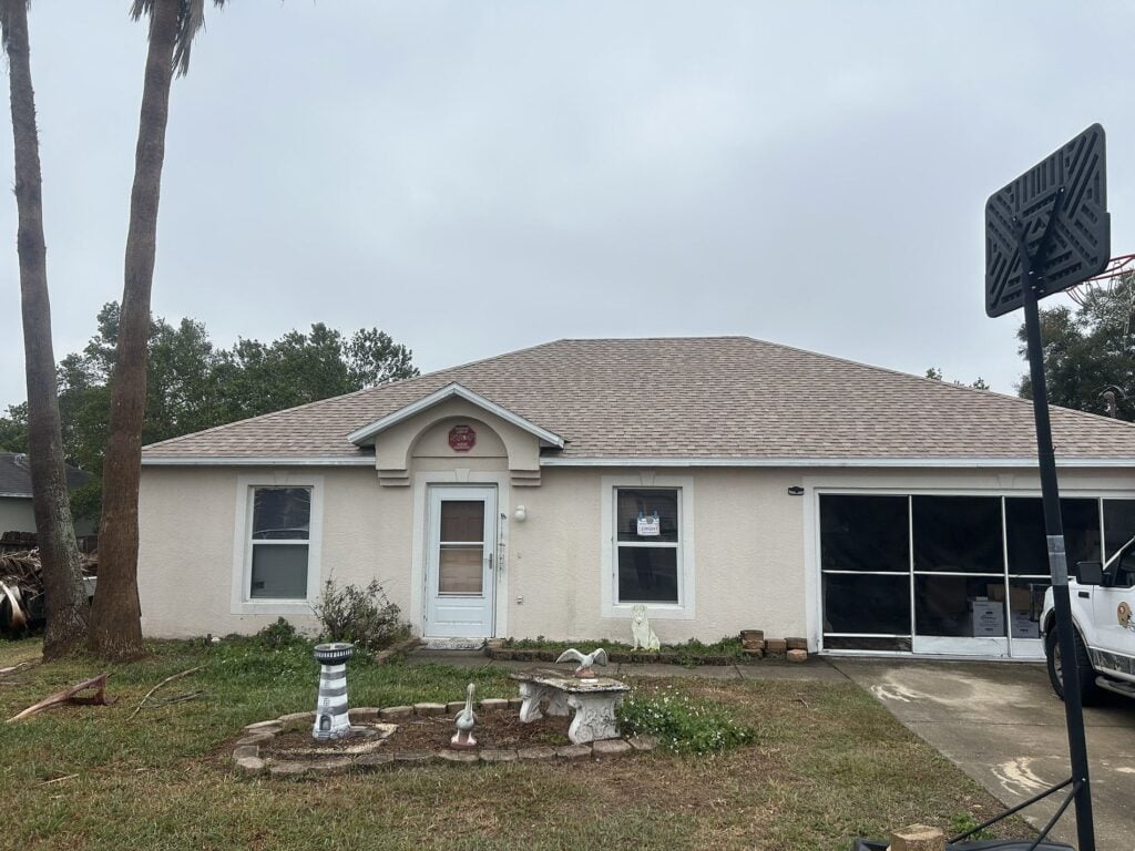 A single-story house with a light-colored exterior, a central front door, two windows, and a screened garage. The yard has birdbaths, statues, and a basketball hoop on the right. Sky is overcast.