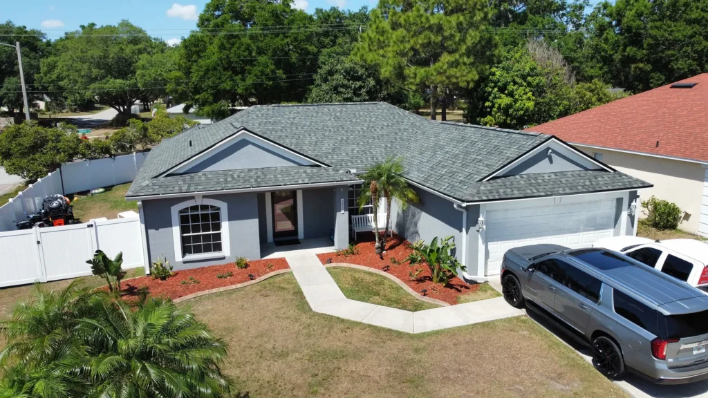 A single-story gray house with a dark gray roofing Tampa shingle roof, manicured front yard, white fenced-in backyard, and two vehicles in the driveway, surrounded by green trees and neighboring houses.