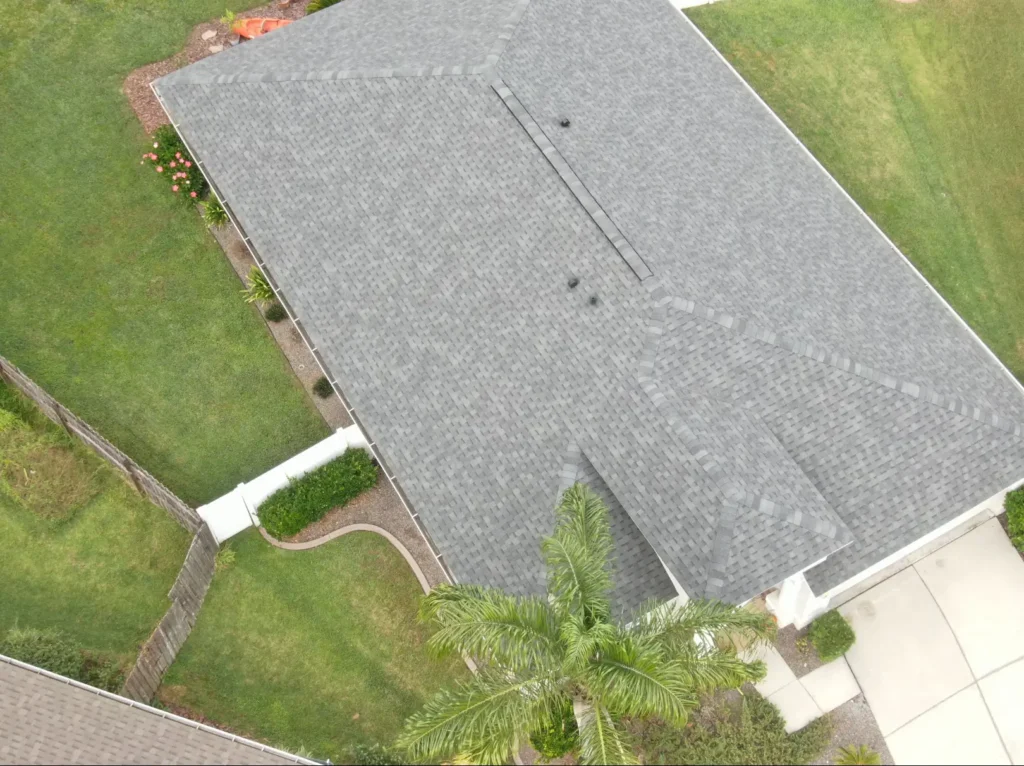 Aerial view of a house with a gray shingled roof by roofing Tampa experts, surrounded by green grass, landscaping, a palm tree, and a white driveway. A fence and flower beds line the yard's edge.
