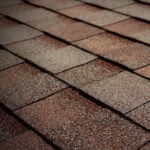 Close-up view of overlapping brown asphalt roof shingles, arranged in neat horizontal rows, displaying a rough, textured surface.