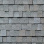 Gray asphalt roof shingles arranged in a staggered, overlapping pattern, creating a textured surface. The shingles have a rough, granular appearance and cover the entire visible area.