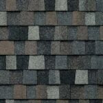 Close-up view of asphalt roof shingles arranged in an overlapping pattern, featuring shades of gray, brown, and black. The textured surface shows slight color variations on each rectangular shingle.