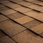 Close-up view of brown asphalt shingles arranged in overlapping rows on a residential roof. The texture of the shingles is visible, showing a mix of light and dark brown tones.