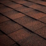 Close-up view of overlapping brown asphalt roof shingles, creating a textured and patterned surface. The lighting highlights the rough granules and the edges of the shingles.