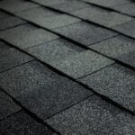 Close-up view of dark asphalt roof shingles arranged in a neat, overlapping pattern. The surface shows a textured, gritty appearance typical of roofing materials.