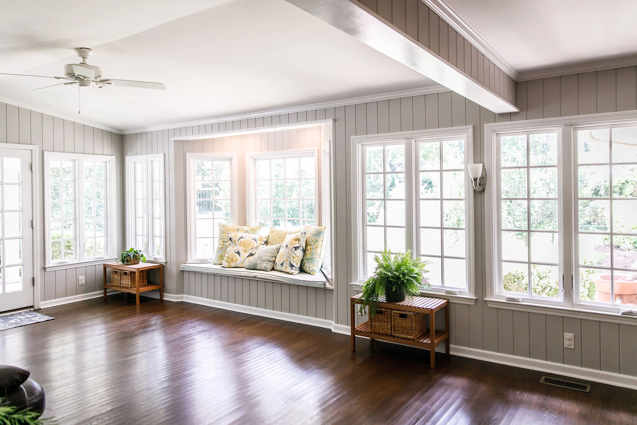 A bright sunroom with hardwood floors, gray paneled walls, large windows, a window seat with floral cushions, and two side tables with potted plants. Natural light fills the airy space, enhanced by quality roofing Tampa homeowners trust.