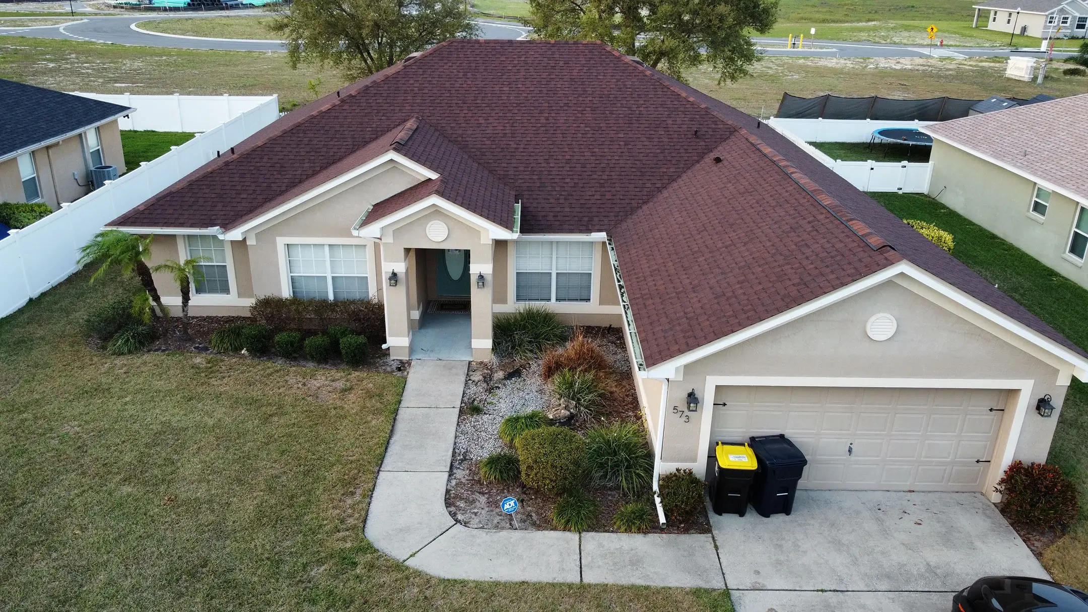 Aerial view of a single-story beige house with a red-brown roof, attached two-car garage, driveway, and well-maintained front yard with shrubs and small trees. Two garbage bins are by the garage.