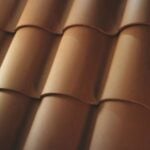 Close-up view of overlapping terracotta roof tiles, arranged in neat, parallel rows, creating a repeating, wavy pattern. The tiles have a smooth, matte finish and a warm, reddish-brown color.