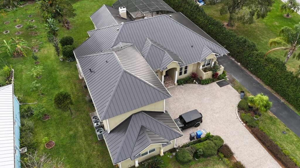 Aerial view of a large beige house with a dark metal roof, surrounded by green lawns and trees. The property includes a paved driveway, a black vehicle, and an enclosed patio or pool area at the back.