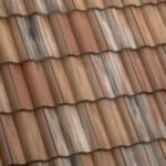 Close-up view of overlapping, wavy terracotta roof tiles with varying shades of brown, red, and tan, creating a textured and weathered appearance.
