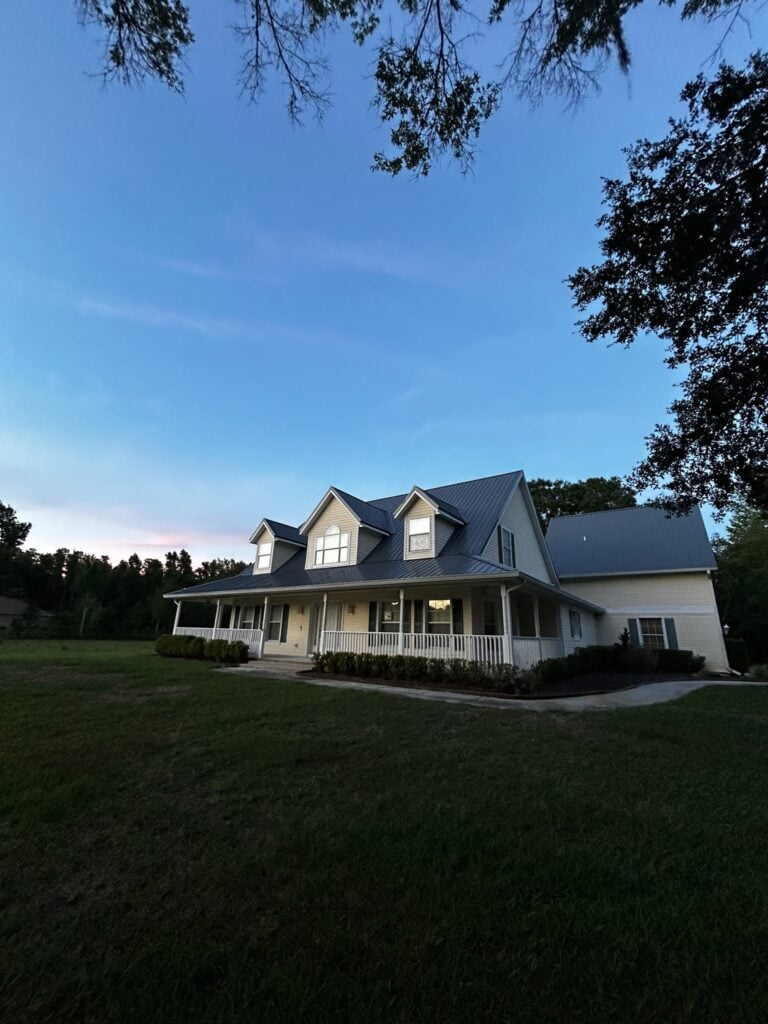 A two-story house with a large front porch, light-colored siding, and a gray metal roof sits on a grassy lawn under a clear evening sky, with trees framing the scene.
