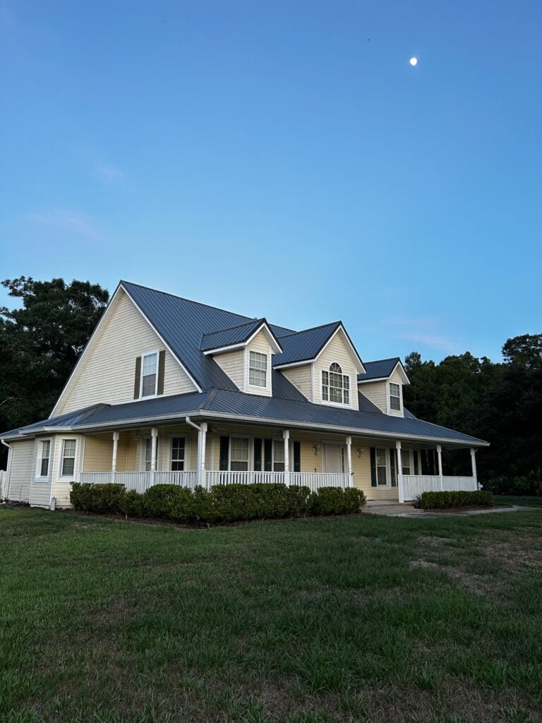 A two-story light yellow house with a large front porch, white trim, and dark gray roof sits on a grassy lawn at dusk, with trees in the background and a small moon visible in the clear blue sky.