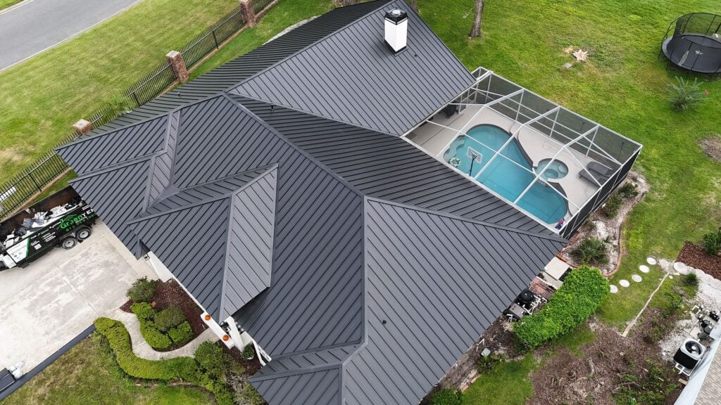 Aerial view of a house with a dark metal roof, a covered backyard pool, green lawn, landscaped garden, driveway, and part of a parked truck visible.