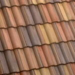 Close-up view of a roof with overlapping wavy tiles in shades of brown, beige, and reddish-orange, arranged in a repeating pattern.