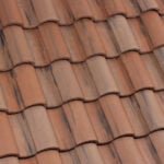 Close-up view of overlapping reddish-brown clay roof tiles with a wavy, textured pattern. The tiles show some darker streaks and natural variation in color, creating a rustic appearance.