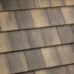 A close-up view of overlapping rectangular roof shingles in shades of brown and gray, showing a textured and weathered surface arranged in a diagonal pattern.