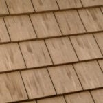 Close-up view of a beige shingle roof with overlapping rectangular tiles arranged in a neat, uniform pattern. The tiles have subtle color variations and streaks, giving them a natural, textured appearance.