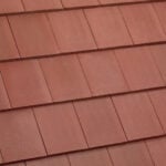Close-up view of overlapping reddish-brown roof tiles arranged in a uniform, horizontal pattern. The tiles have a smooth, matte texture and are neatly aligned.