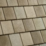 Close-up view of a roof covered with rectangular, overlapping beige shingles arranged in neat horizontal rows. The shingles have a slightly textured surface, showing some variation in color.