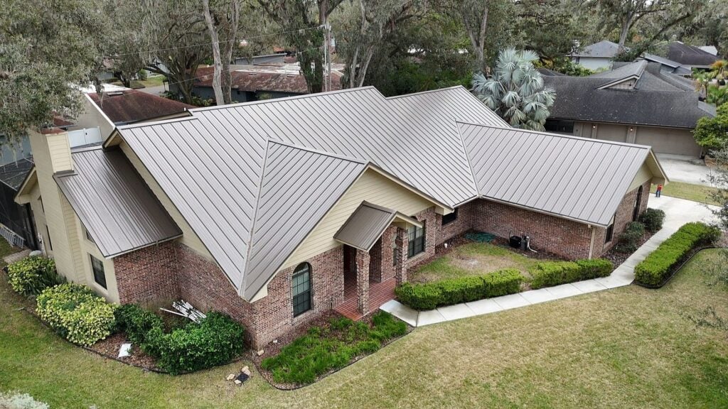 Aerial view of a single-story brick house with a light brown metal roof, surrounded by grass, shrubs, and trees. A concrete walkway borders the front and side of the house. Neighboring houses are visible in the background.