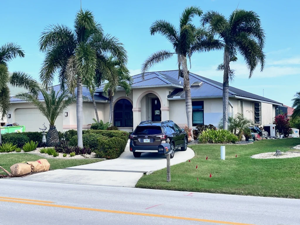 Single-story house with a dark roof, surrounded by palm trees and landscaped shrubs. A black SUV is parked in the driveway. The house has arched windows and a manicured front lawn next to a residential street.