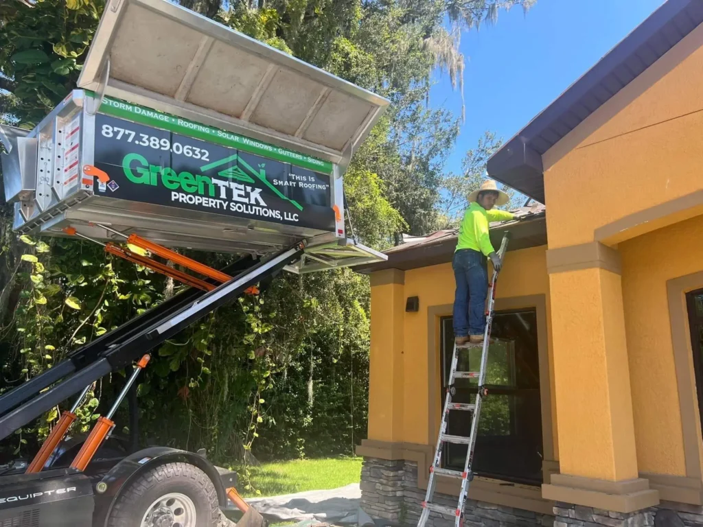 A worker in a neon shirt and hard hat stands on a ladder beside a house, inspecting the roof near a raised trailer labeled "GreenTek Property Solutions, LLC" under sunny weather with trees in the background.