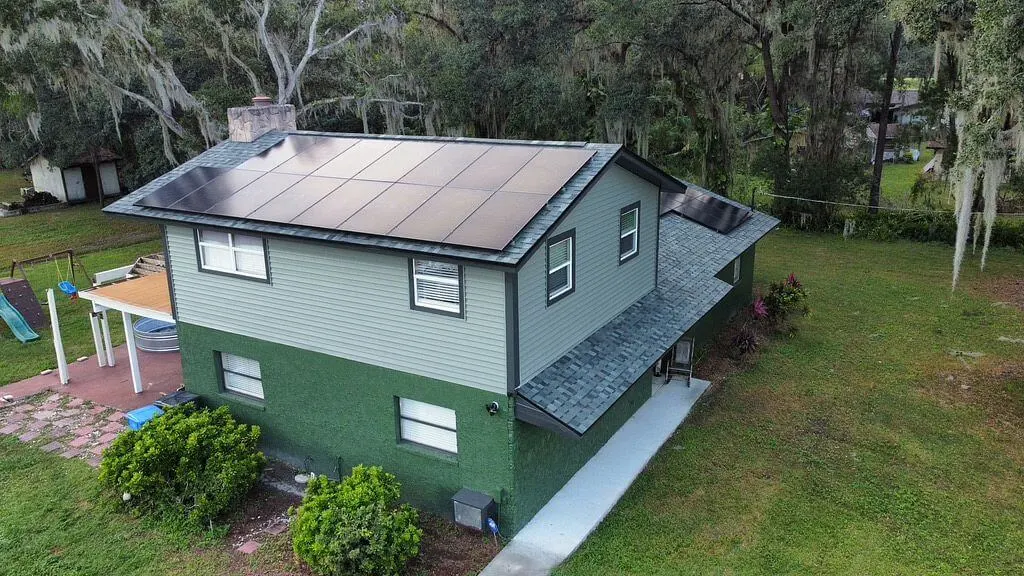 A two-story house with green siding and a gray roof features solar panels installed on both main roof sections, surrounded by grass, trees, and a play area in the backyard.