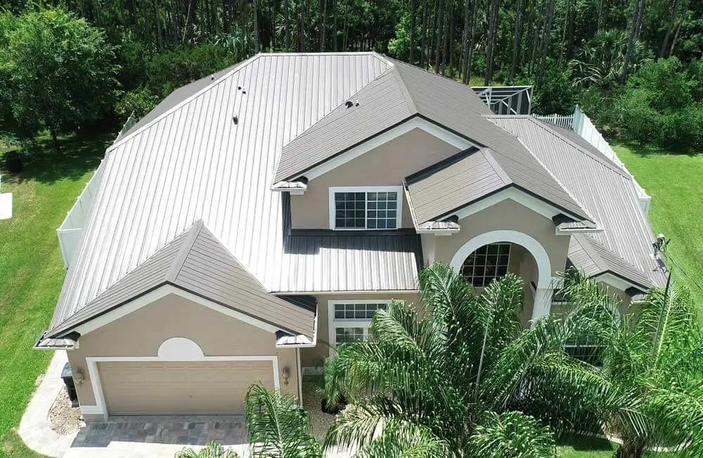 Aerial view of a two-story house with a beige exterior, large white-trimmed windows, and a brown metal roof, surrounded by green lawn and palm trees, with a garage on the left side.