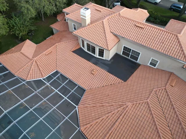Aerial view of a large house with multiple sections and orange clay tile roofing. A porch area and a screened pool enclosure are visible, surrounded by trees and greenery.
