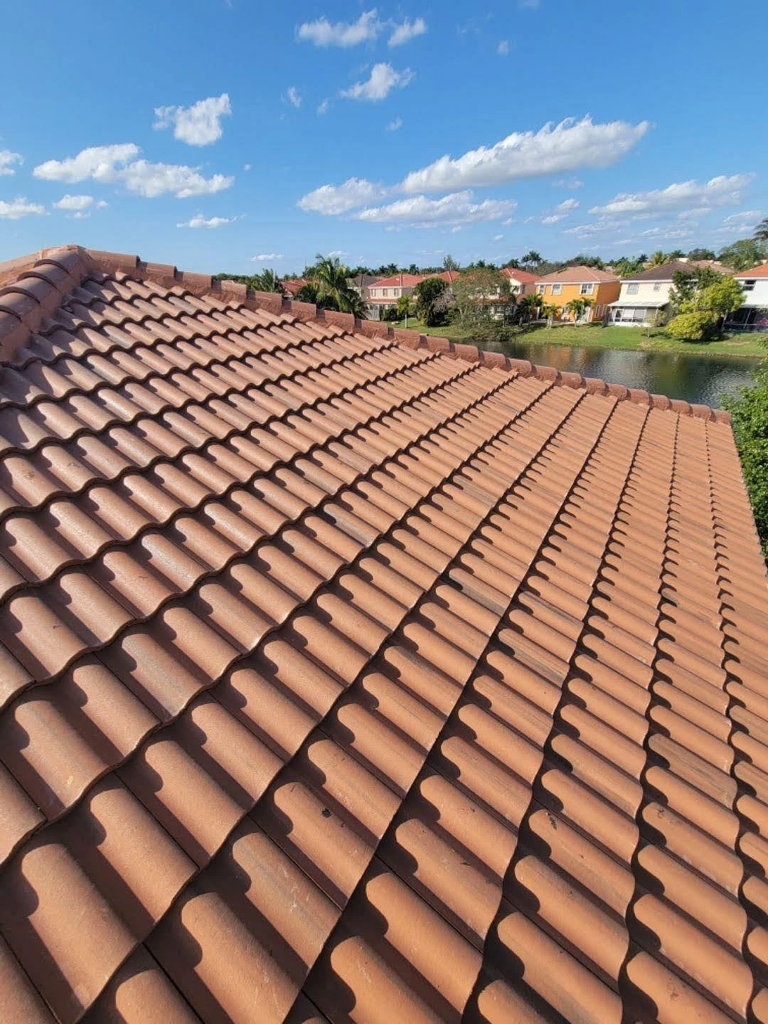 A close-up view of a terracotta-tiled roof under a blue sky with scattered clouds, overlooking a suburban neighborhood with houses, greenery, and a body of water in the background.