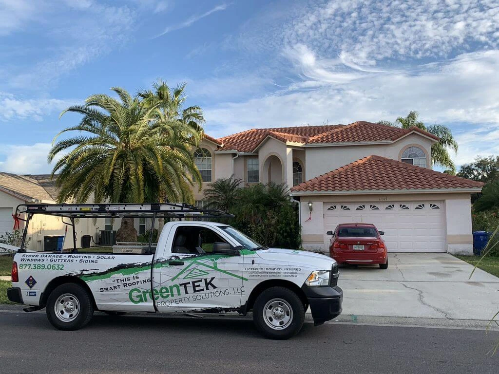 A white GreenTEK Property Solutions pickup truck is parked in front of a suburban house with a red tile roof, palm trees, and a red car in the driveway under a blue sky with scattered clouds.