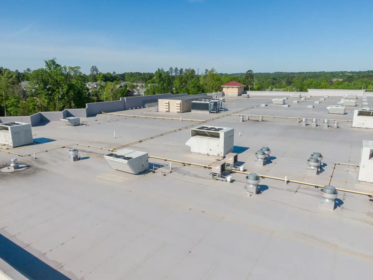 A large flat commercial rooftop, ideal for flat roof replacement, with multiple HVAC units, vents, and ducts, surrounded by trees and buildings under a clear blue sky.