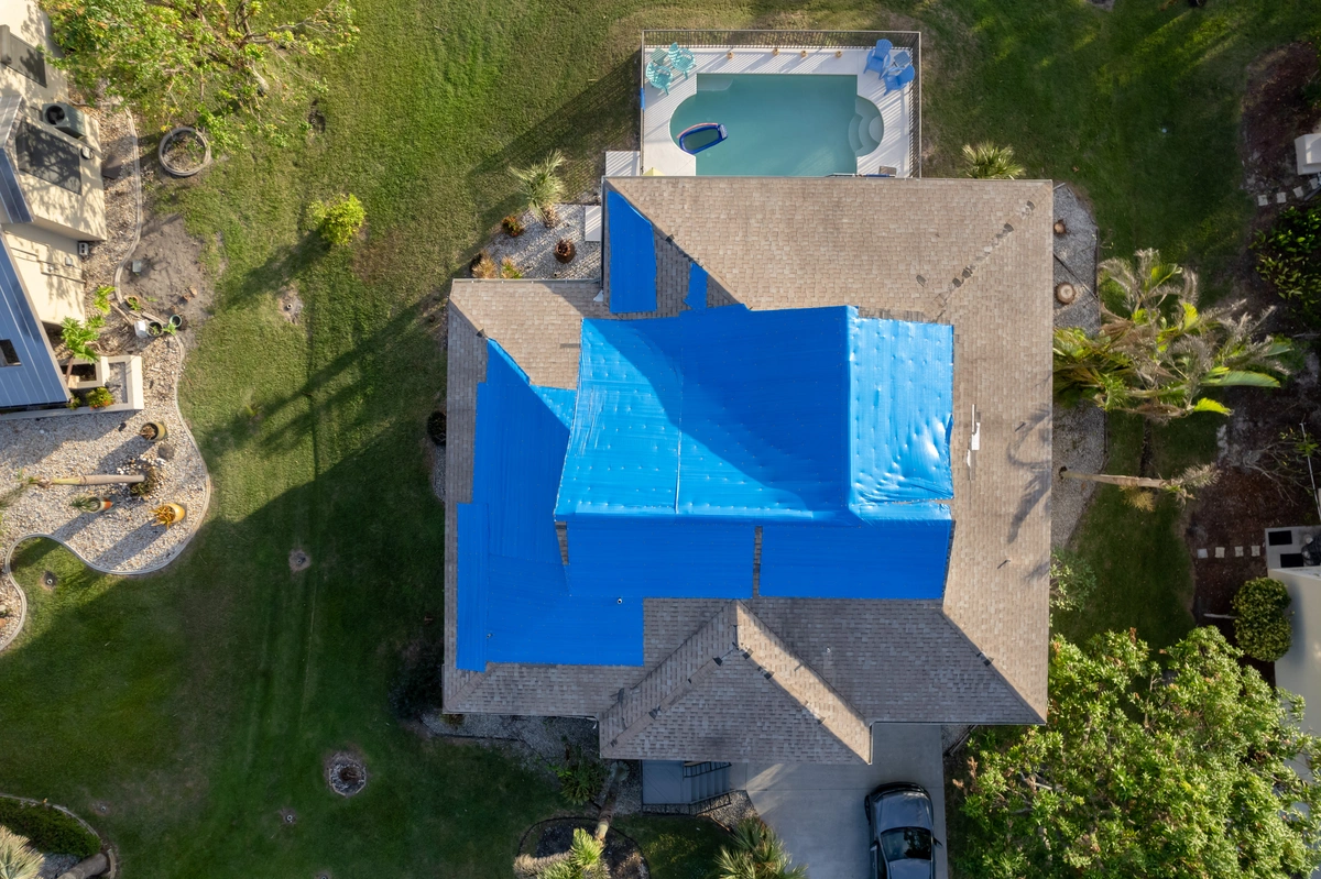 Aerial view of a house with blue tarps covering parts of its roof, suggesting recent diy roof repair, surrounded by a yard with trees and a swimming pool in the backyard. A car is parked in the driveway.
