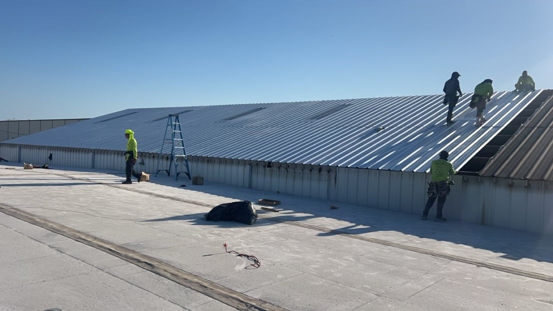 Construction workers in safety gear install or repair a large metal roof on a building under a clear blue sky. A ladder stands nearby on the roof, and tools and materials are scattered around.