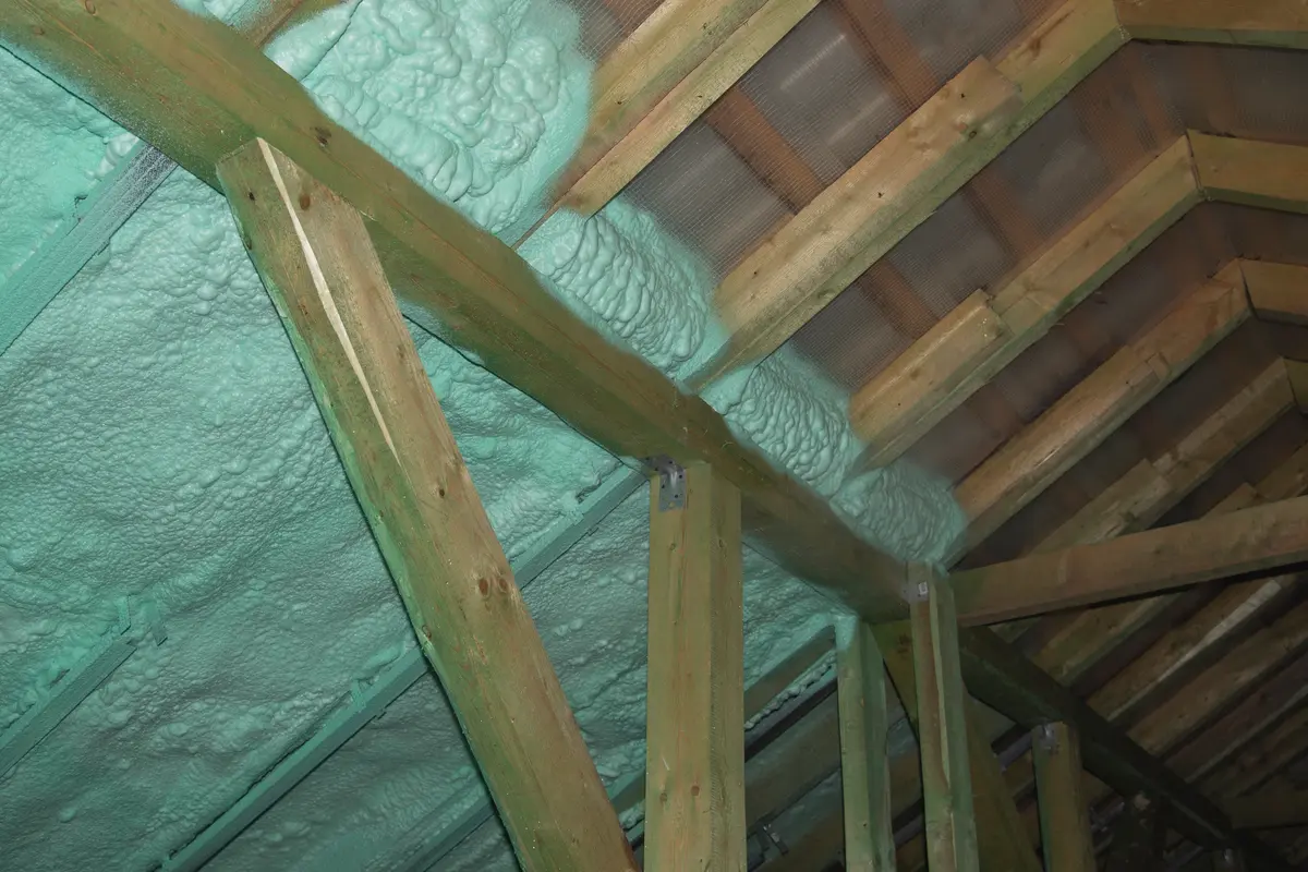Spray foam roofing insulation is applied to the underside of a roof between wooden rafters in an unfinished attic, with greenish foam filling gaps for improved thermal efficiency.