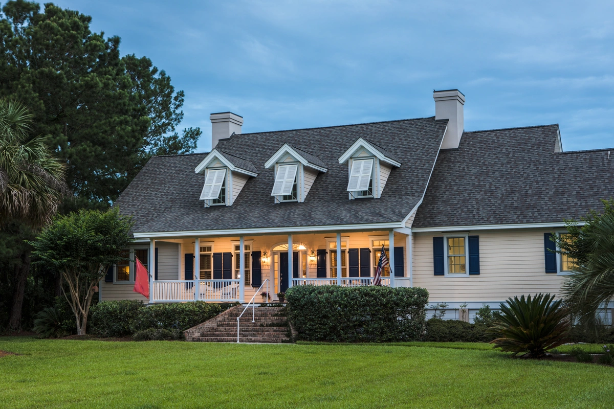 A large, light-colored house with a steep, dark gray roof featuring popular GAF shingle colors, three dormer windows, a front porch with lights on, and a well-maintained lawn surrounded by trees and landscaping.