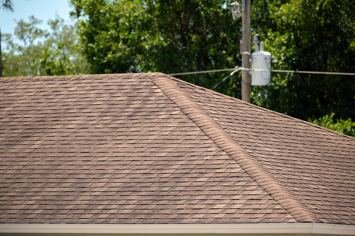 A close-up view of a brown shingle roof on a building highlights its condition, suggesting that emergency roof repair may be needed, with trees and a utility pole in the background under a clear blue sky.