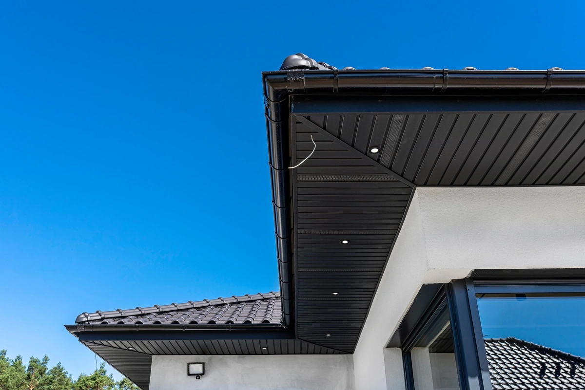 Close-up view of a modern house roof with black tiles, black gutter system, and sleek soffit panels with recessed lights under the eaves of a house. A clear blue sky is in the background.